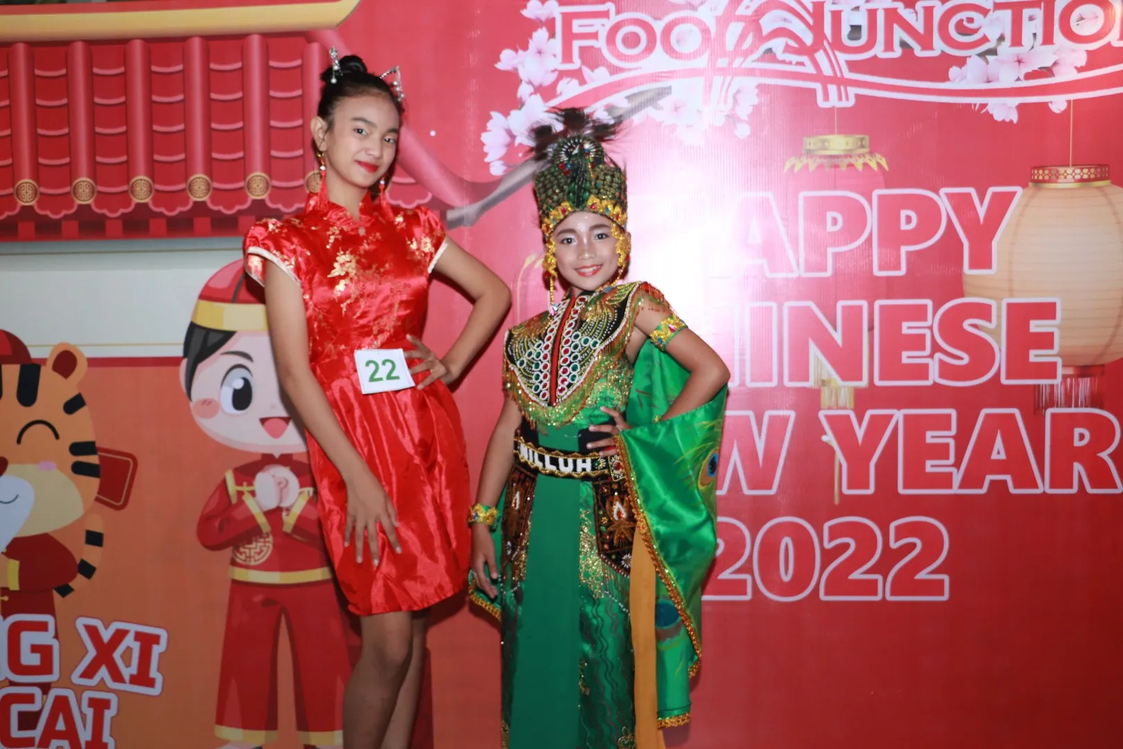 Two young girls dressed in traditional and festive costumes posing in front of a red Chinese New Year backdrop